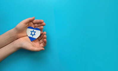children's hands hold a heart cut out of paper with a drawn Israeli flag on a blue background....