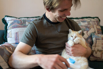 young adult man grooming a yellow cat with a brush, pet owner brushing a cute kitten in the living...