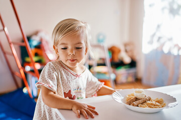 Little girl stands near the table in front of a plate of breakfast and looks away