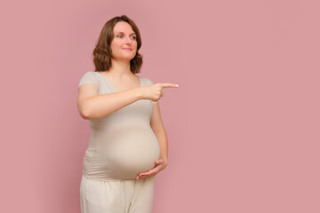 A finger gesture towards a pregnant woman on a studio pink background. Pregnancy in a woman with a belly with a hand pointing to the side, copy space