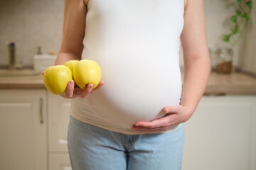 A pregnant woman holds apples in her hands while standing in her home kitchen. Pregnancy and proper...