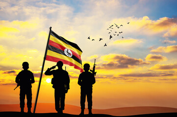 Silhouettes of soldiers with the Uganda flag stand against the background of a sunset or sunrise....