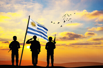 Silhouettes of soldiers with the Uruguay flag stand against the background of a sunset or sunrise....