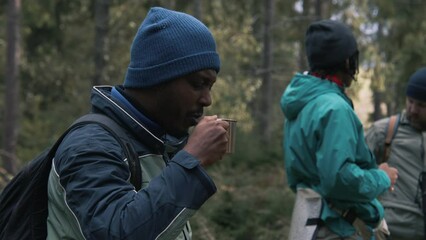 African American tourist keeps cup in hand and shares hot tea with female traveler. Group of multiethnic hiking buddies stopped to rest in forest camping. Nature discovery and tourism. Slow motion.