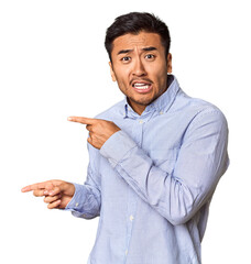 Young Chinese man in studio background shocked pointing with index fingers to a copy space.