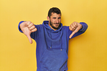 Young Hispanic man on yellow background showing thumb down and expressing dislike.