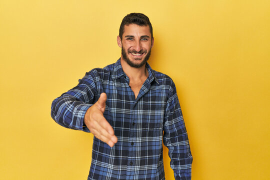 Young Hispanic man on yellow background stretching hand at camera in greeting gesture.