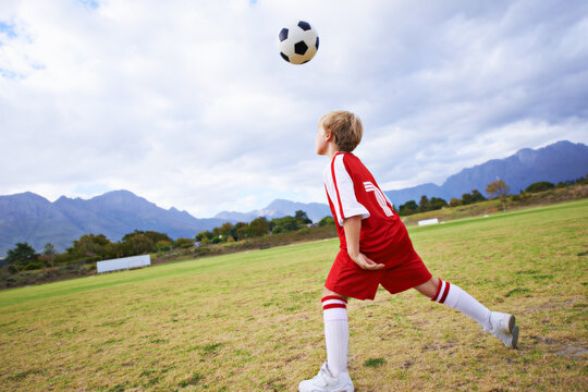 Kid, soccer ball and playing on green grass for sports, training or practice with clouds and blue sky. Young football player or athlete ready for kick off, game or match on outdoor field in nature