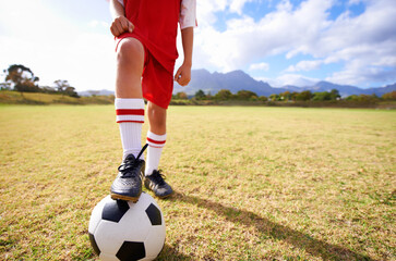 Child, soccer ball and legs on green grass for sports, training or practice with clouds and blue...