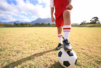 Child, soccer ball and legs on green field for sports, training or practice with clouds and blue...
