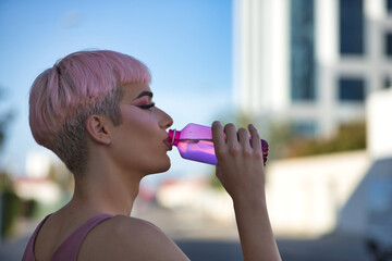 Portrait of young, attractive, gay, heavily makeup man with pink hair and top, drinking water from...