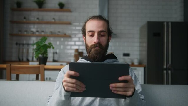 Young bearded hair bun man sitting on sofa at home with kitchen background, playing excited concentrated engrossed in interesting mobil game with tablet computer