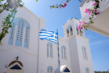 View of the greek flag waving in the air next to a bougainvillea and a whitewashed church in the...