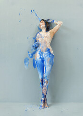 sexy naked proud beauty woman, hand up, in blue and white color painted, standing at the wall, shows her painted body. Creative, abstract expressive bodypainting art