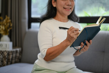 Smiling woman 50s years old wearing casual clothes writing her diary while relaxing at home.