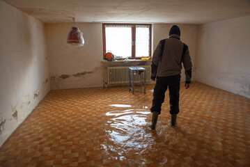 Man walks with rubber boots in a flooded basement room during flood - obrazy, fototapety, plakaty