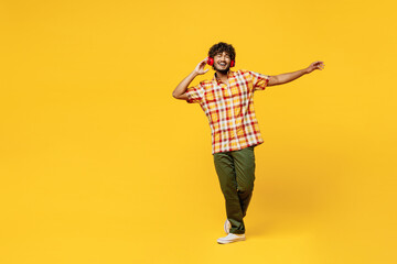 Full body young cheerful fun happy cool Indian man he wearing shirt casual clothes dancing listen to music in headphones isolated on plain yellow color background studio portrait. Lifestyle concept. - obrazy, fototapety, plakaty