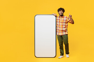 Full body young Indian man wears shirt casual clothes big huge blank screen mobile cell phone smartphone with mockup area point finger up isolated on plain yellow color background. Lifestyle concept.