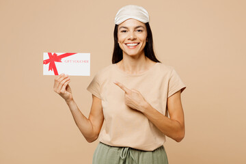 Young calm Latin woman wear pyjamas jam sleep eye mask rest relax at home hold point on gift coupon voucher card for store isolated on plain pastel beige background studio Good mood night nap concept