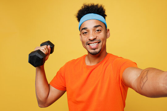Young fitness trainer sporty man sportsman wear orange t-shirt hold dumbbell do selfie shot mobile cell phone training in home gym isolated on plain yellow background. Workout sport fit abs concept.