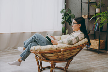 Full body sideways young smart woman wear casual clothes glasses sits on armchair read book novel...