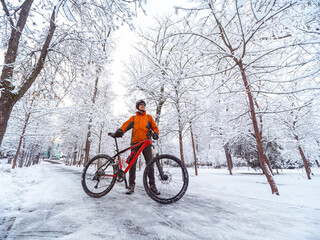 A man with a bicycle in an orange jacket and a bicycle helmet is in a winter city among snow...