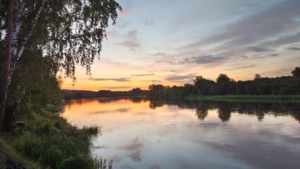 On a summer evening the sun among the trees has dipped below the horizon. The colorful cloudy sky is reflected in the calm river water. Trees stand on the grassy banks, and calamus grows in the water