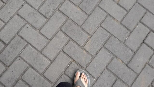 Video Or Footage Of A Pair Of Feet Wearing Sandals Walking Down A Cobbled Street Paved Concrete Blocks In A Gray Zigzag Pattern. Paving Blocks are Made From A Mixture of Cement, Water and Aggregate