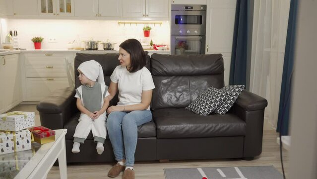 3-year-old child and his mother are sitting on sofa waiting for Santa Claus, little boy is scared