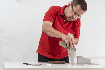 Latin Man in red shirt using saw on furniture piece, deeply involved in DIY crafting.