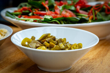 A close up of a bowl of jalapenos ready to eat, with salad defocused behind