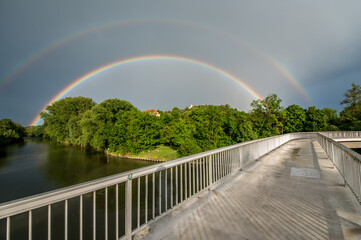 Doppelter Regenbogen vor  dunklen Wolken an Fluss mit Fussgängersteg und Flussufer mit Bäumen und...