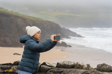 Woman with a knit hat and jacket trying to take a photo from a cliff with a lonely beach in the...