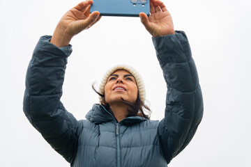 Latin woman with knit hat and coat taking a photo with her cell phone seen from below with cloudy...