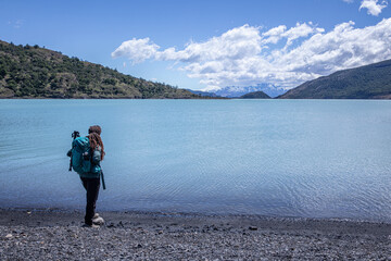 A female hiker standing on a stone beach looking away to Patagonia landscape with mountains, clouds, glaciers, and a blue lake