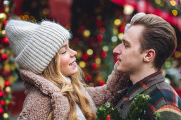 Beautiful young loving couple, boyfriend and girlfriend having fun on a christmas market wearing warm clothes. Outdoors, winter time, snowy weather. Tangerines in string bag, fir tree branches