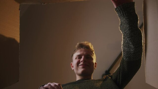 Close-up inside of carton box bottom view, young blonde man opens parcel looks at delivered goods excited, smiling feels satisfied 