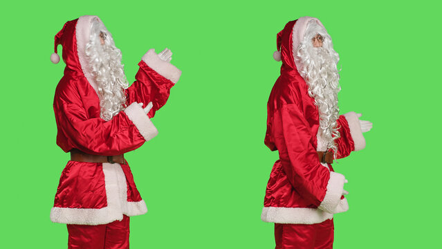 Father christmas advertising something, pointing aside while he stands against greenscreen backdrop with stanta claus festive costume. Man dressed like saint nick, xmas marketing campaign.