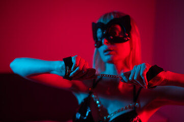 Fototapeta na wymiar Blonde girl in a leather mask and handcuffs in the bedroom in red blue neon light. Role-playing games.