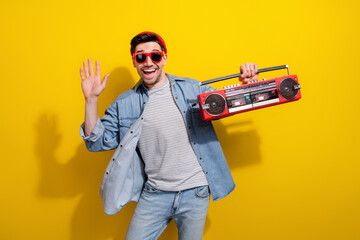 Photo of funky friendly man dressed jeans shirt dark spectacles waving arm hi listening boom box songs isolated yellow color background
