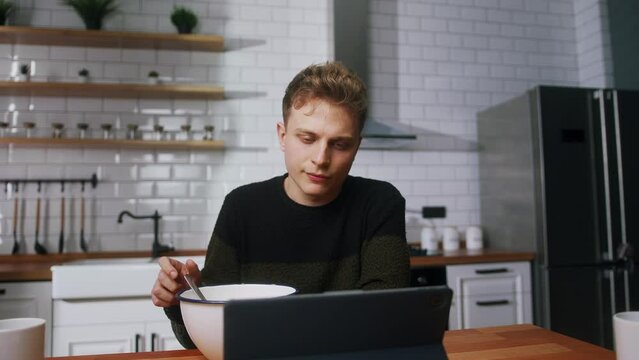 Young bored man reluctant to eat pushing a bowl full of cereal while sitting in the kitchen watching a video on a tablet computer