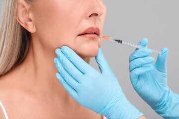 Doctor giving lips injection to senior woman on light grey background, closeup. Cosmetic surgery