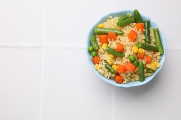 Delicious bulgur with vegetables in bowl on white tiled table, top view. Space for text