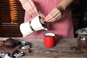Woman pouring aromatic coffee from moka pot into cup at wooden table indoors, closeup
