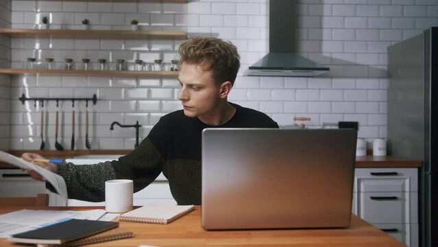 Young handsome university student sitting in the kitchen at home, looking laptop computer, studying, taking notes. Male student researching, study at home