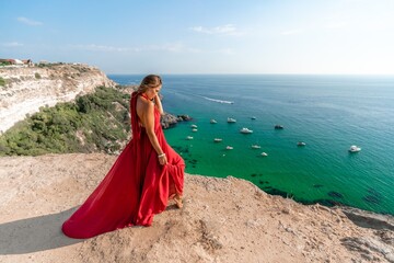 Fototapeta na wymiar Woman sea red dress yachts. A beautiful woman in a red dress poses on a cliff overlooking the sea on a sunny day. Boats and yachts dot the background.