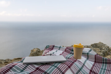 Laptop coffee on blanket with ocean view. Illustrating serene outdoor laptop use. Freelancer enjoying their time outdoors while working or browsing the internet.