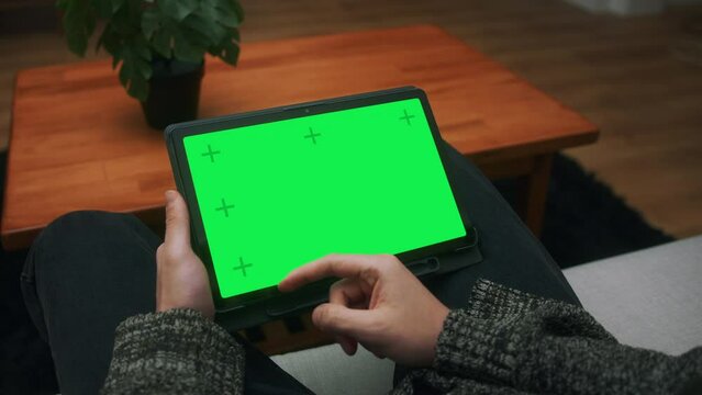 Young Man Scrolling And Tapping Center On Tablet With Green Screen Mock Up Display. Male Sitting On Sofa, Relaxing At Home. Close-up Over The Shoulder Shot