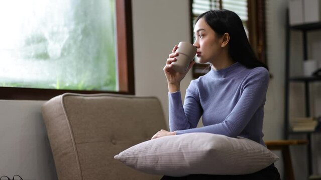 Beautiful Asian woman at home drinking coffee while relaxing on the sofa in the living room.