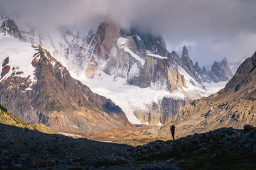 A tiny woman standing alone in the Agostini campsite surrounded by mountain range in the morning with Mt.Cerro torre as background (Patagonia)